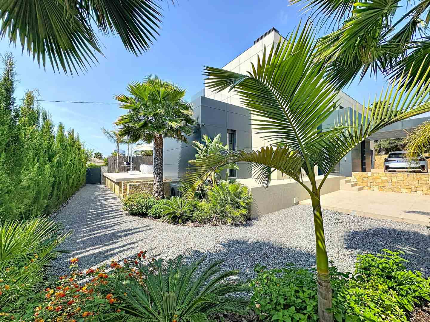 Vista exterior de la villa con fachada minimalista gris. Jardín paisajístico con caminos de grava, palmeras y vegetación tropical, enmarcado por un seto alto bajo un cielo azul claro.