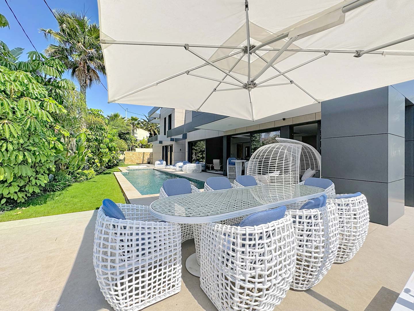 Zona de comedor exterior con mesa de cristal y sillas de diseño en blanco y cojines azules, bajo una gran sombrilla. Al fondo, piscina privada y exuberante jardín tropical junto a la moderna fachada de la villa.