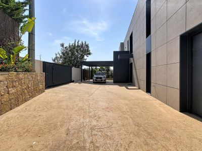 Amplia entrada con pavimento de hormigón texturizado, muro de piedra natural y portón automático oscuro. Destaca la fachada moderna de paneles grises y el carport integrado.