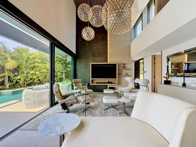 Amplio salón de doble altura con grandes ventanales que conectan directamente con la piscina privada y el jardín tropical. Destacan los tres candelabros esféricos y el revestimiento de mármol en la pared.