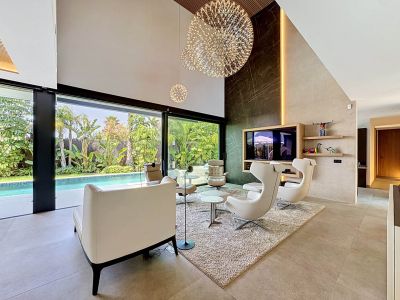 Amplio salón de doble altura con pared de acento de efecto mármol oscuro, elegantes lámparas esféricas y grandes puertas de cristal que se abren a una piscina privada y jardín tropical.