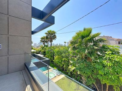Balcón moderno con fachada de paneles grises y barandilla de cristal, revelando jacuzzis exteriores, exuberante vegetación tropical y el skyline de Calpe al fondo.