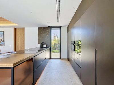 Cocina de diseño con mobiliario oscuro sin tiradores y electrodomésticos Miele integrados. La isla central cuenta con encimera negra, placa y fregadero, complementada por una pared de piedra veteada y salida al jardín.