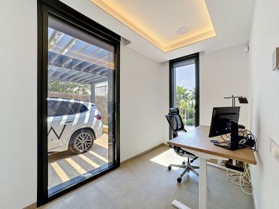 Despacho moderno con escritorio de roble claro, silla ergonómica y luz natural de una puerta corredera que muestra un garaje exterior y una ventana con vistas a jardines tropicales.
