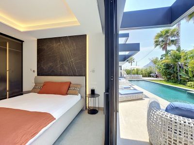 Dormitorio con acceso directo a la piscina privada y jardín tropical. Destaca una pared de acento efecto mármol oscuro con iluminación integrada y mobiliario exterior moderno.