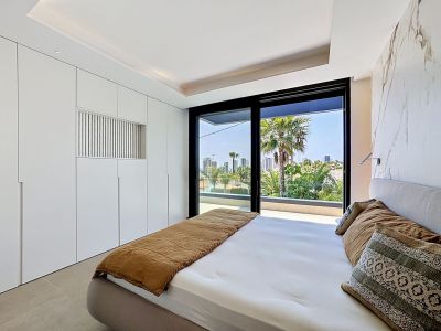 Dormitorio moderno con armarios empotrados blancos y pared de acento efecto mármol. Grandes puertas de cristal dan a una terraza privada con vistas a palmeras y al horizonte urbano.