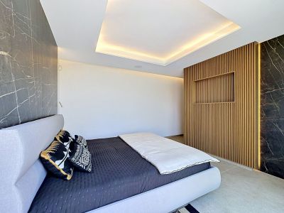 Dormitorio moderno con cama tapizada en gris claro, paneles de pared efecto mármol oscuro y una pared central de lamas de madera con iluminación integrada y estante empotrado. El techo presenta iluminación perimetral.