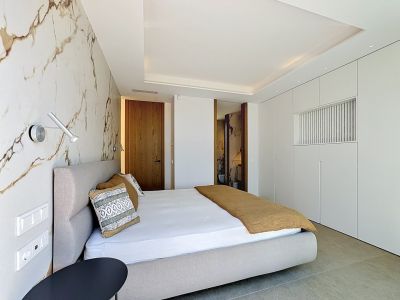 Dormitorio principal con pared de acento de mármol, cama tapizada y armarios blancos integrados. Una puerta de madera conduce al baño en suite.