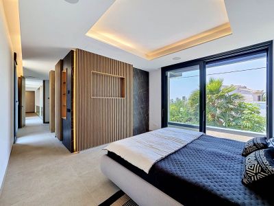 Dormitorio principal con una pared de lamas de madera iluminadas y panel de mármol oscuro. Grandes ventanales ofrecen vistas a la vegetación y arquitectura local, complementado por iluminación cenital.