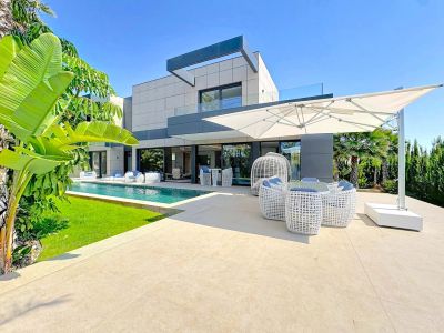 El exterior de la villa presenta una piscina rectangular, terraza de baldosas claras con mobiliario lounge blanco moderno y una gran sombrilla voladiza, rodeado de paisajismo tropical.