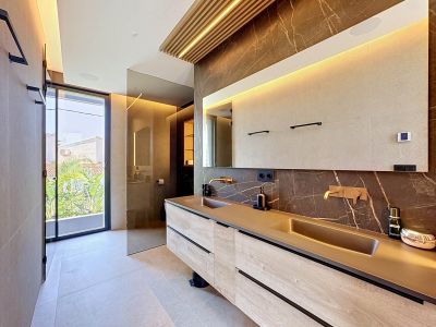 Elegante baño con doble lavabo integrado, grifería de oro rosa y pared de mármol oscuro veteado. Un detalle de techo de madera con luz indirecta y acceso a la terraza exterior.