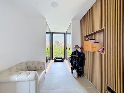 Elegante zona de vestidor con pared de lamas de madera y estantería integrada, un sofá clásico de cuero capitoné y grandes puertas de cristal hacia jardines.