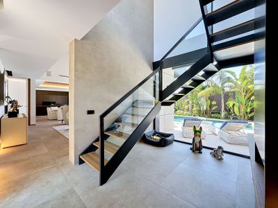 Escalera moderna de peldaños abiertos con barandilla de cristal y metal negro, conectando interiores diáfanos y vistas a la piscina privada con jardín tropical.