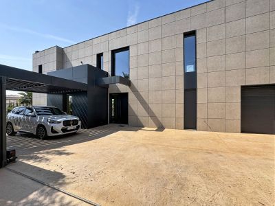 Fachada minimalista de la villa con revestimiento de paneles claros y entrada en antracita. El porche de lamas metálicas protege un vehículo, junto a un amplio acceso pavimentado y garaje.
