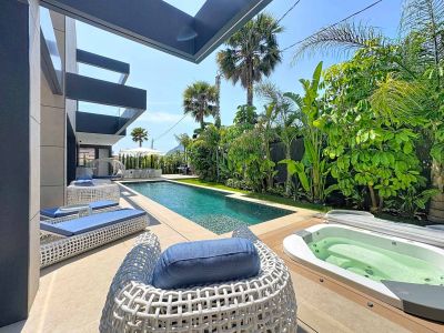 La zona exterior moderna ofrece una piscina rectangular, un jacuzzi integrado en tarima de madera y mobiliario lounge blanco con cojines azules. Jardines tropicales exuberantes aportan privacidad.