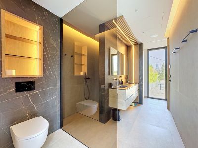 Moderno baño con paredes de efecto mármol oscuro, tocador flotante de roble con doble lavabo y pared de lamas de madera iluminada. Mampara de cristal separa la ducha a ras de suelo y el inodoro suspendido.