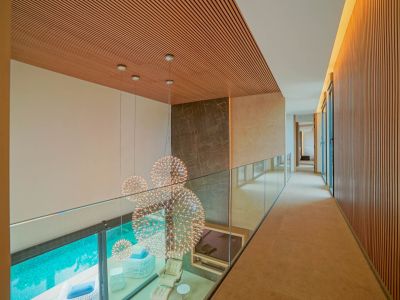 Pasillo superior con barandilla de cristal y techo de lamas de madera, ofreciendo vistas a un salón de doble altura con lámparas esféricas y acceso a la piscina.