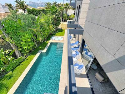 Piscina rectangular con jacuzzi integrado, rodeada de exuberantes jardines tropicales y césped. Balcones modernos con mobiliario de exterior y fachada de baldosas grises.
