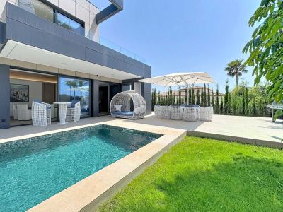 Terraza exterior con piscina de mosaico azul, césped y mobiliario de diseño blanco. La moderna fachada de la villa se abre a este espacio de ocio con zona de comedor bajo sombrilla y barbacoa.
