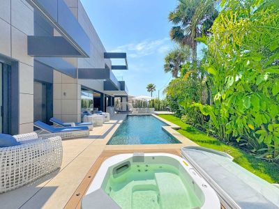 Terraza exterior con piscina rectangular y jacuzzi integrado. Muebles de diseño en blanco y azul complementan la fachada moderna de la villa y el exuberante jardín tropical.