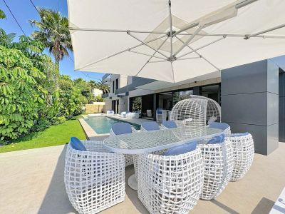 Zona de comedor exterior con mesa de cristal y sillas de diseño en blanco y cojines azules, bajo una gran sombrilla. Al fondo, piscina privada y exuberante jardín tropical junto a la moderna fachada de la villa.