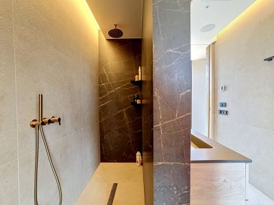 Zona de ducha moderna con azulejos efecto mármol oscuro veteado y paredes claras texturizadas. Dispone de ducha de lluvia y de mano en bronce, e iluminación LED perimetral. Se aprecia parte de un mueble de lavabo.