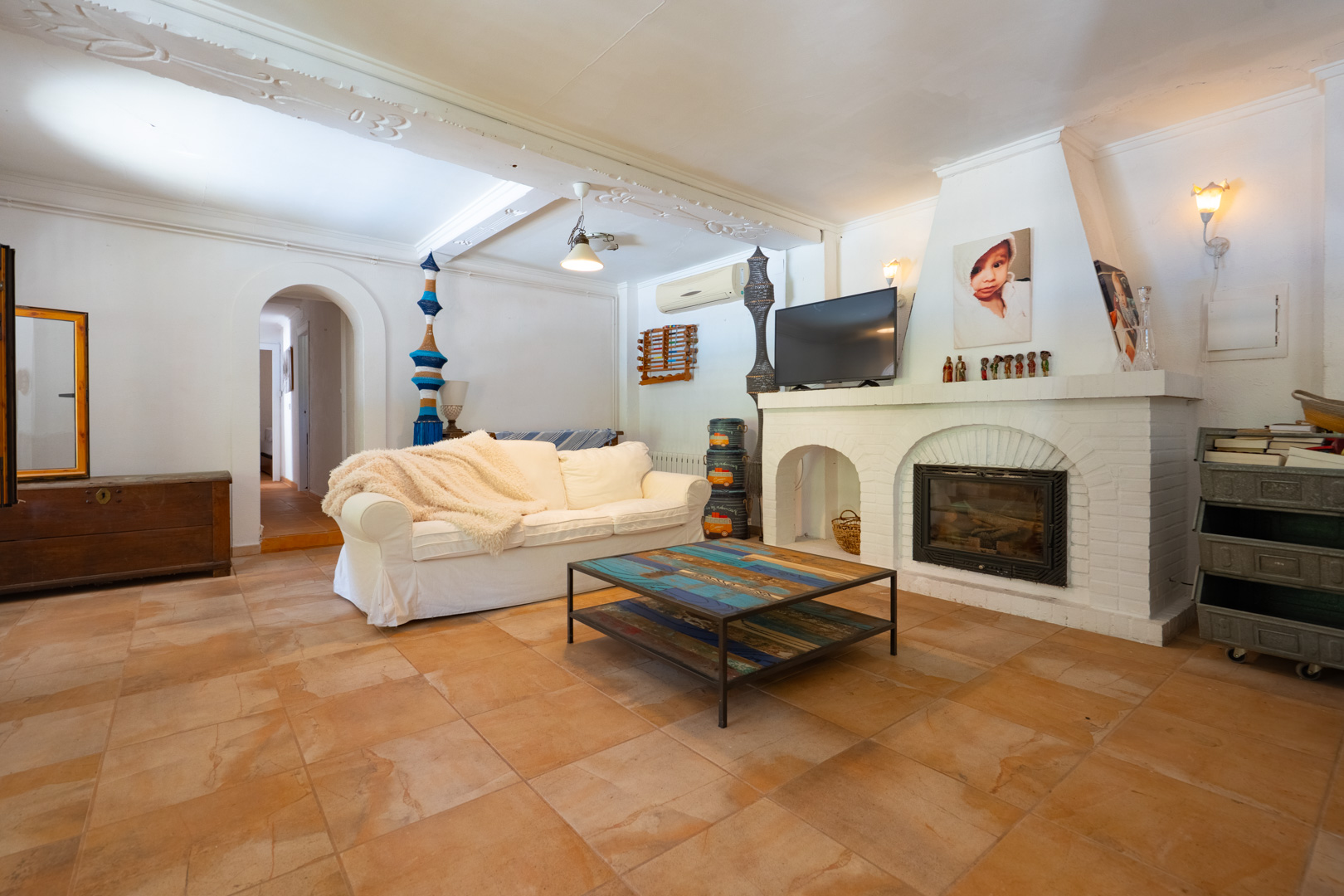 Acogedor salón con chimenea de ladrillo blanco y televisión, un cómodo sofá y una mesa de centro única con tapa de madera reciclada sobre baldosas de terracota. Acogedor salón con chimenea de ladrillo blanco y televisión, un cómodo sofá y una mesa de centro única con tapa de madera reciclada sobre baldosas de terracota.