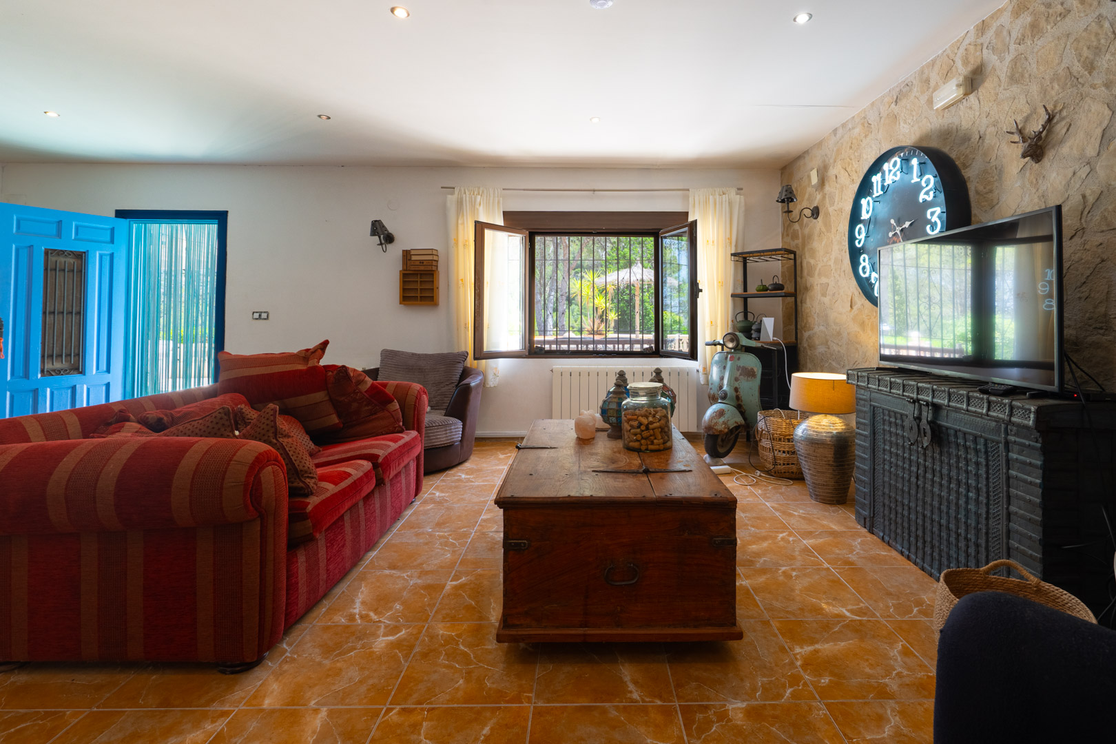 Acogedora sala de estar con sofá rojo a rayas, mesa de centro de baúl de madera rústica y pared de piedra con reloj azul grande y TV. Acogedora sala de estar con sofá rojo a rayas, mesa de centro de baúl de madera rústica y pared de piedra con reloj azul grande y TV.