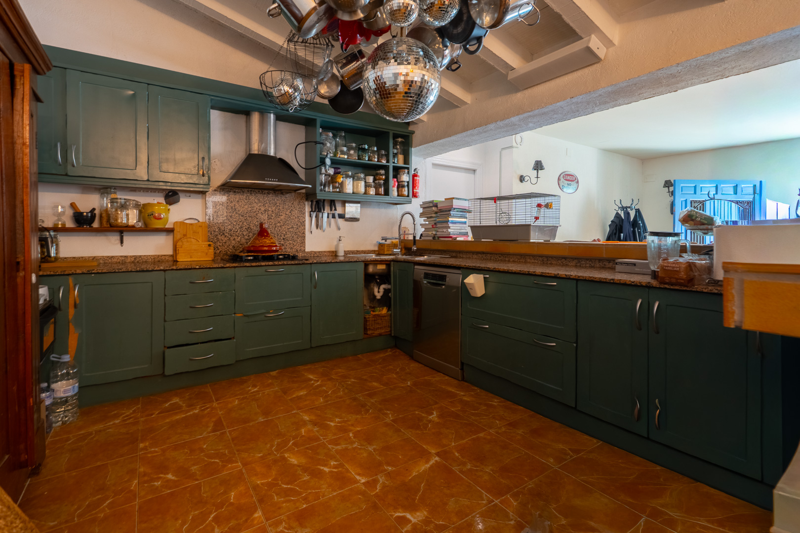 Amplia cocina con muebles en verde oscuro, encimeras de granito y suelo de baldosas de terracota. Su diseño semiabierto conecta con una estancia adyacente, destacando vigas vistas y decoración ecléctica. Amplia cocina con muebles en verde oscuro, encimeras de granito y suelo de baldosas de terracota. Su diseño semiabierto conecta con una estancia adyacente, destacando vigas vistas y decoración ecléctica.