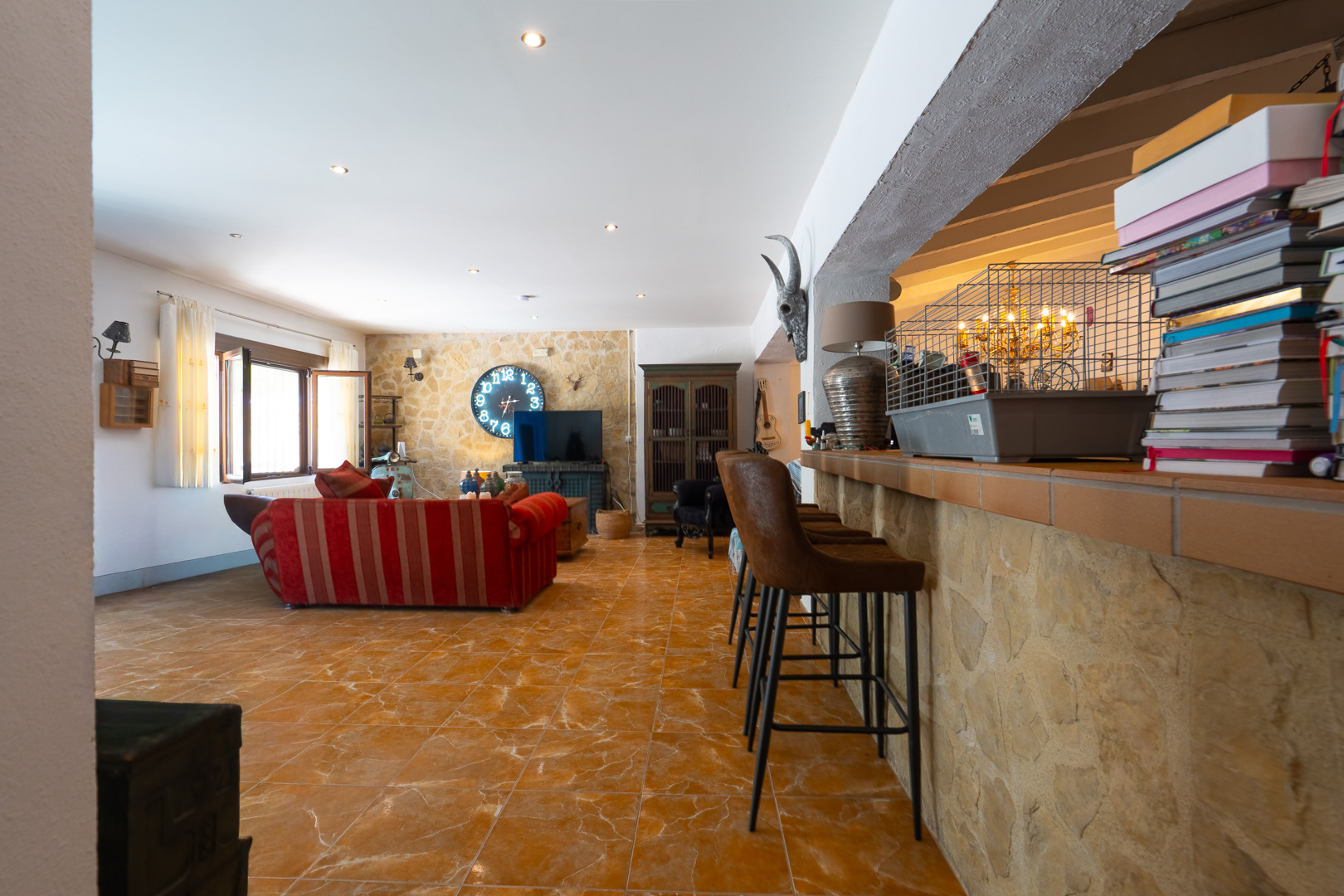 Amplia sala de estar con barra rústica revestida de piedra y tres taburetes, conectando con un salón que tiene sofá rojo, suelo de baldosas y un gran reloj circular en la pared de piedra. Amplia sala de estar con barra rústica revestida de piedra y tres taburetes, conectando con un salón que tiene sofá rojo, suelo de baldosas y un gran reloj circular en la pared de piedra.