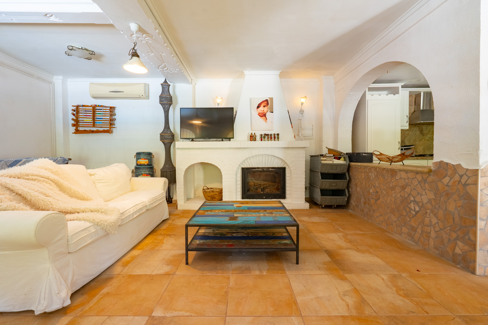 Amplio salón con chimenea de ladrillo blanco y TV integrada, mesa de centro de madera multicolor única y suelo de baldosas de terracota. Un arco conecta con la cocina semiabierta. Amplio salón con chimenea de ladrillo blanco y TV integrada, mesa de centro de madera multicolor única y suelo de baldosas de terracota. Un arco conecta con la cocina semiabierta.
