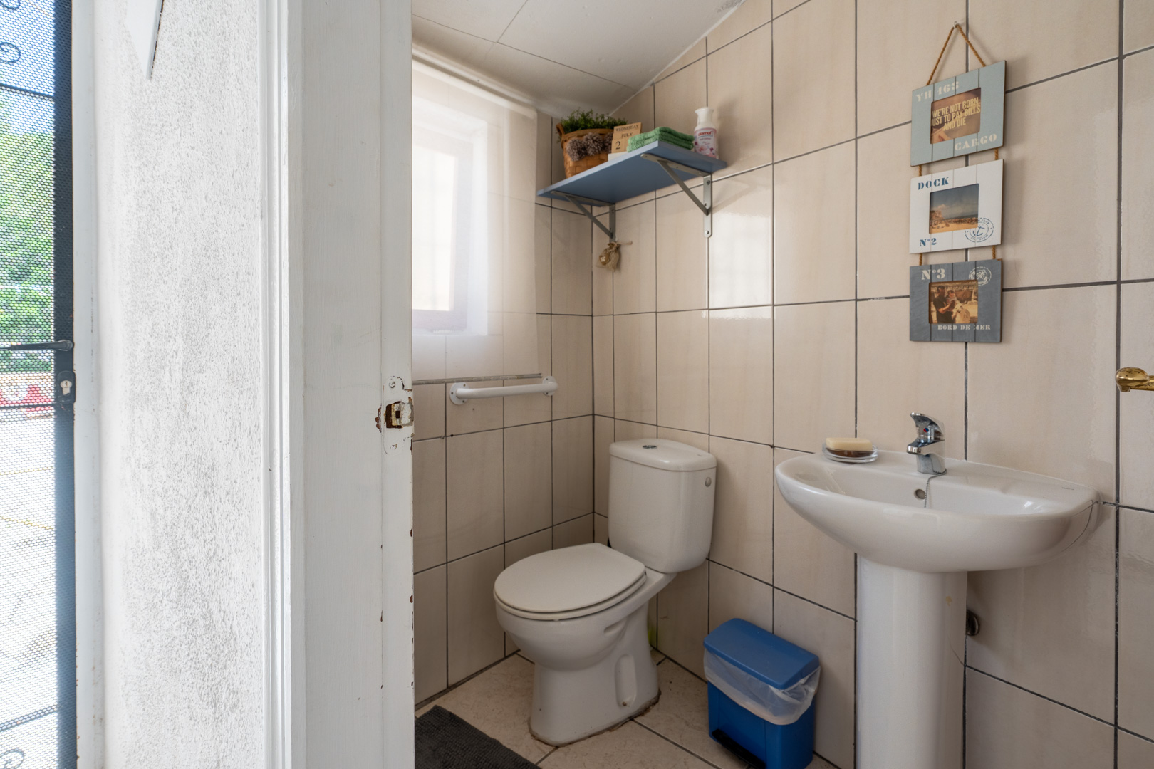 Baño auxiliar compacto con azulejos beige, inodoro y lavabo de pedestal blancos, luz natural de una ventana pequeña y estante decorativo. Baño auxiliar compacto con azulejos beige, inodoro y lavabo de pedestal blancos, luz natural de una ventana pequeña y estante decorativo.