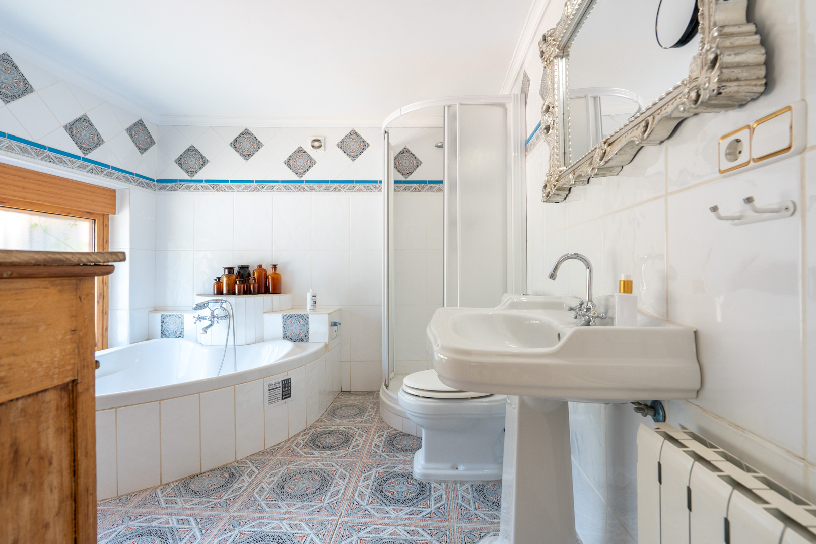 Baño con bañera esquinera, lavabo de pedestal y ducha. Presenta azulejos blancos con cenefas decorativas y suelo hidráulico de patrones geométricos. Baño con bañera esquinera, lavabo de pedestal y ducha. Presenta azulejos blancos con cenefas decorativas y suelo hidráulico de patrones geométricos.