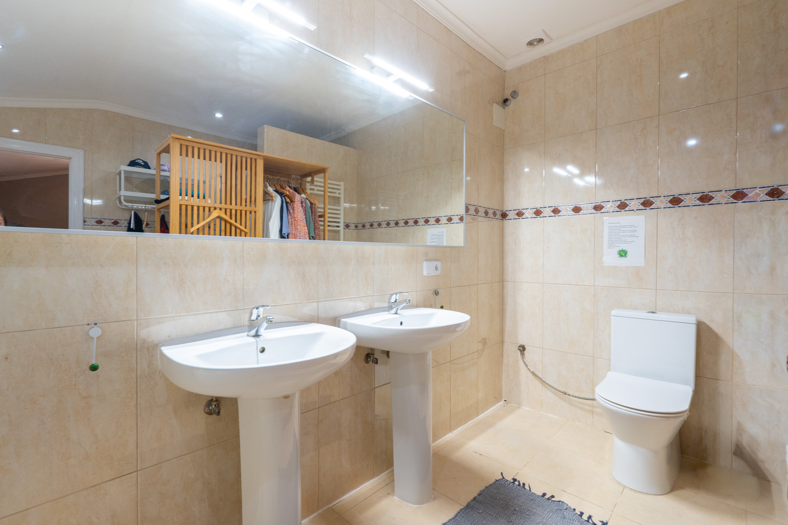 Baño con dos lavabos de pedestal, paredes alicatadas en tonos beige con cenefa decorativa y un amplio espejo que refleja un perchero de madera. Baño con dos lavabos de pedestal, paredes alicatadas en tonos beige con cenefa decorativa y un amplio espejo que refleja un perchero de madera.