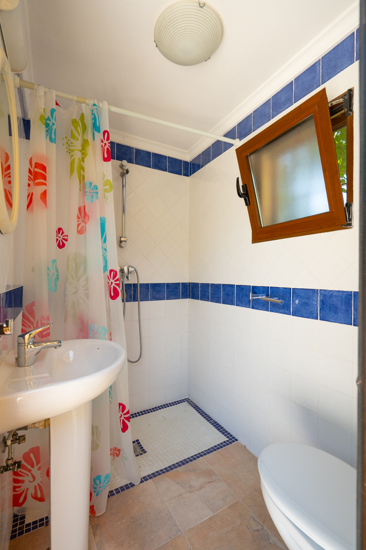 Baño con lavabo de pedestal blanco, ducha con cortina floral colorida y paredes de azulejos blancos con cenefa azul. Una ventana de madera abierta aporta luz natural. Baño con lavabo de pedestal blanco, ducha con cortina floral colorida y paredes de azulejos blancos con cenefa azul. Una ventana de madera abierta aporta luz natural.