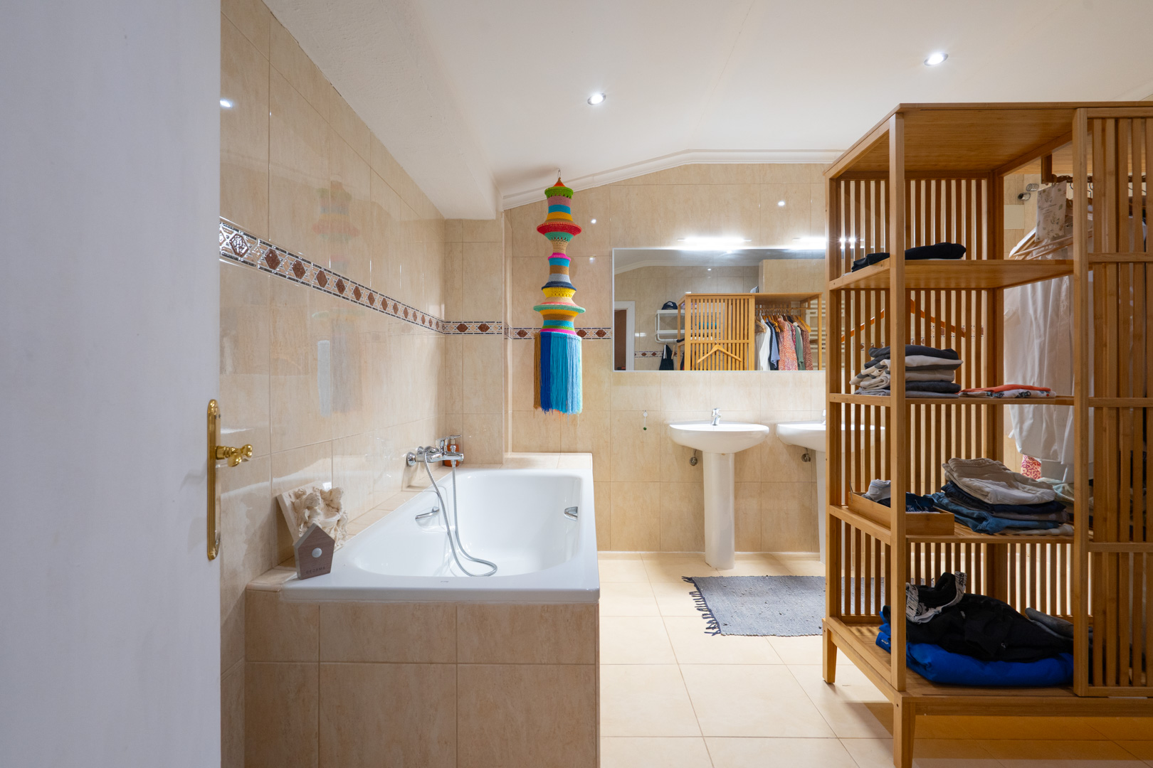 Baño con paredes de azulejos beige claro y cenefa decorativa, bañera y dos lavabos de pedestal. Un gran espejo refleja un mueble de estanterías de madera, iluminado por focos empotrados. Baño con paredes de azulejos beige claro y cenefa decorativa, bañera y dos lavabos de pedestal. Un gran espejo refleja un mueble de estanterías de madera, iluminado por focos empotrados.