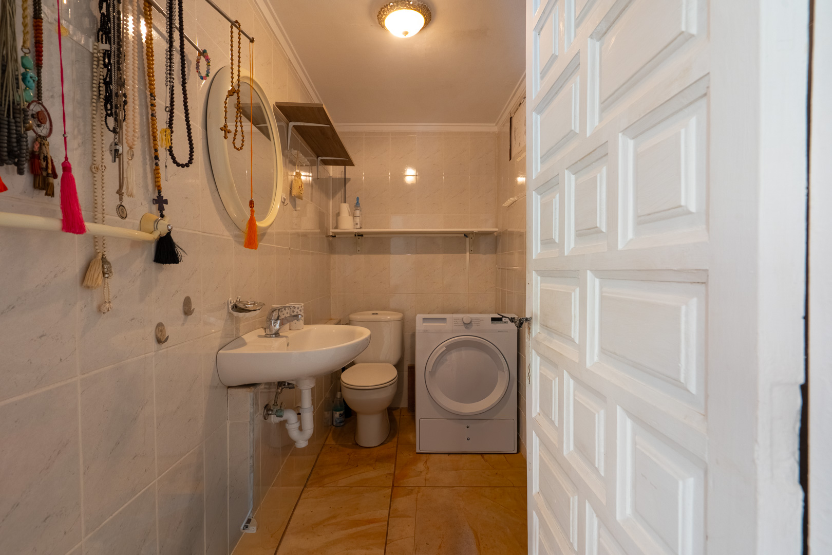 Baño funcional con azulejos claros, suelo de terracota y puerta panelada blanca. Incluye lavabo, inodoro y lavadora, optimizando el espacio. Baño funcional con azulejos claros, suelo de terracota y puerta panelada blanca. Incluye lavabo, inodoro y lavadora, optimizando el espacio.