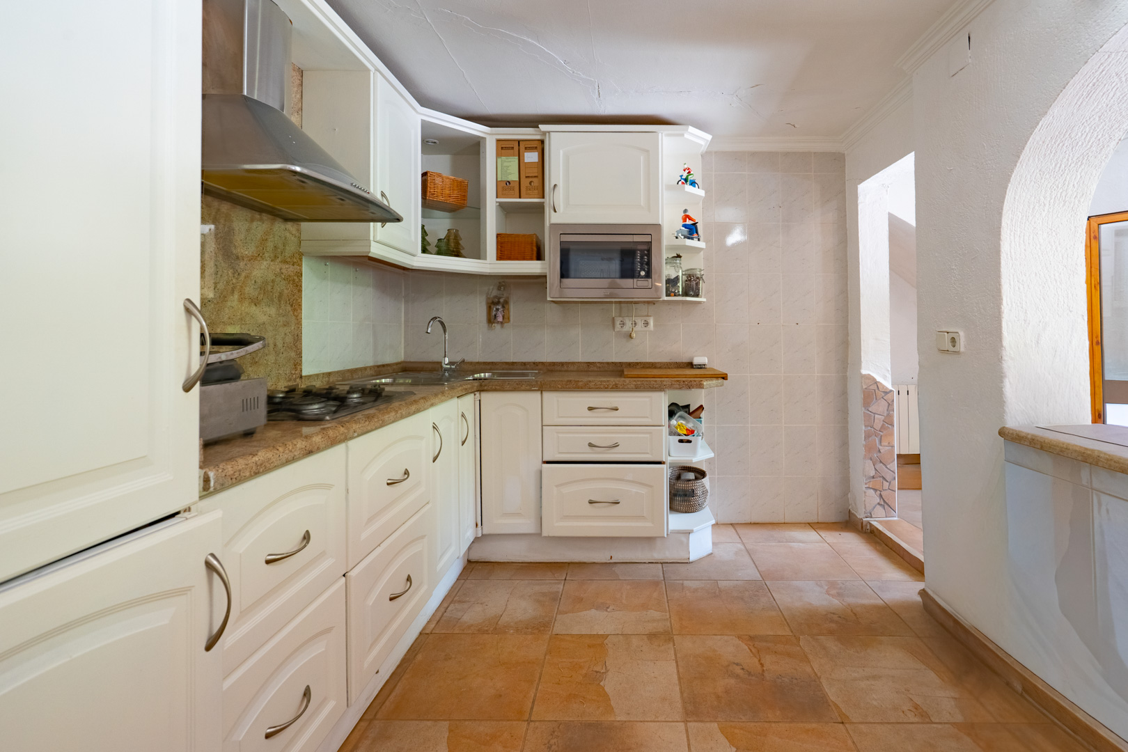 Cocina semiabierta con muebles blancos, encimeras de granito y electrodomésticos integrados. El suelo de baldosas terracota complementa las paredes claras y el arco de acceso. Cocina semiabierta con muebles blancos, encimeras de granito y electrodomésticos integrados. El suelo de baldosas terracota complementa las paredes claras y el arco de acceso.