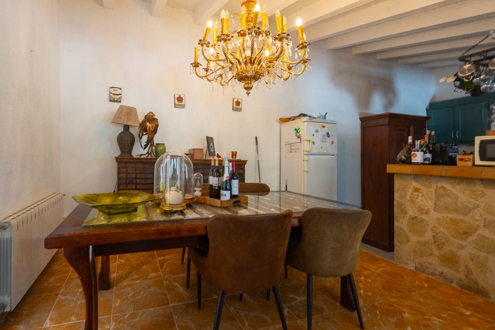 Comedor con mesa de madera oscura y cristal, sillas tapizadas marrones, y una lámpara de araña dorada. Conecta con una barra de cocina revestida de piedra y vigas vistas. Comedor con mesa de madera oscura y cristal, sillas tapizadas marrones, y una lámpara de araña dorada. Conecta con una barra de cocina revestida de piedra y vigas vistas.