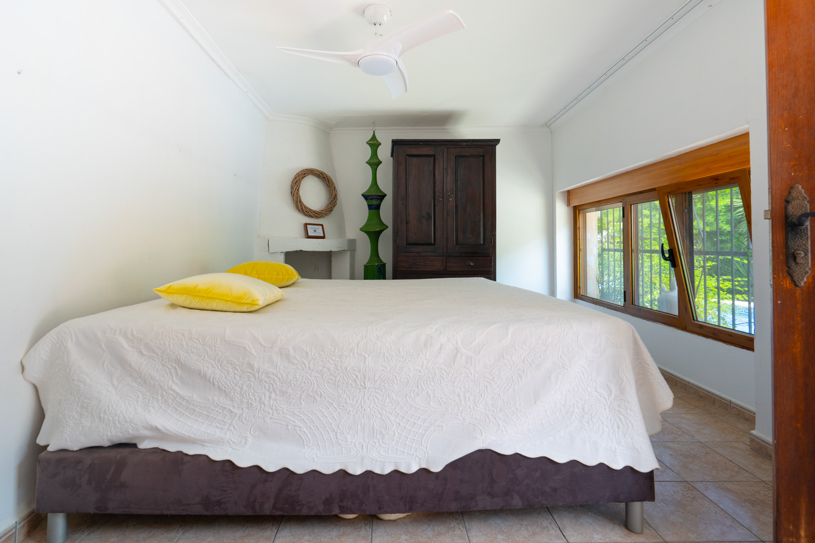 Dormitorio con cama de colcha blanca estampada, armario de madera oscura y ventana de madera que da a la vegetación. Incluye ventilador de techo y suelo de baldosas. Dormitorio con cama de colcha blanca estampada, armario de madera oscura y ventana de madera que da a la vegetación. Incluye ventilador de techo y suelo de baldosas.
