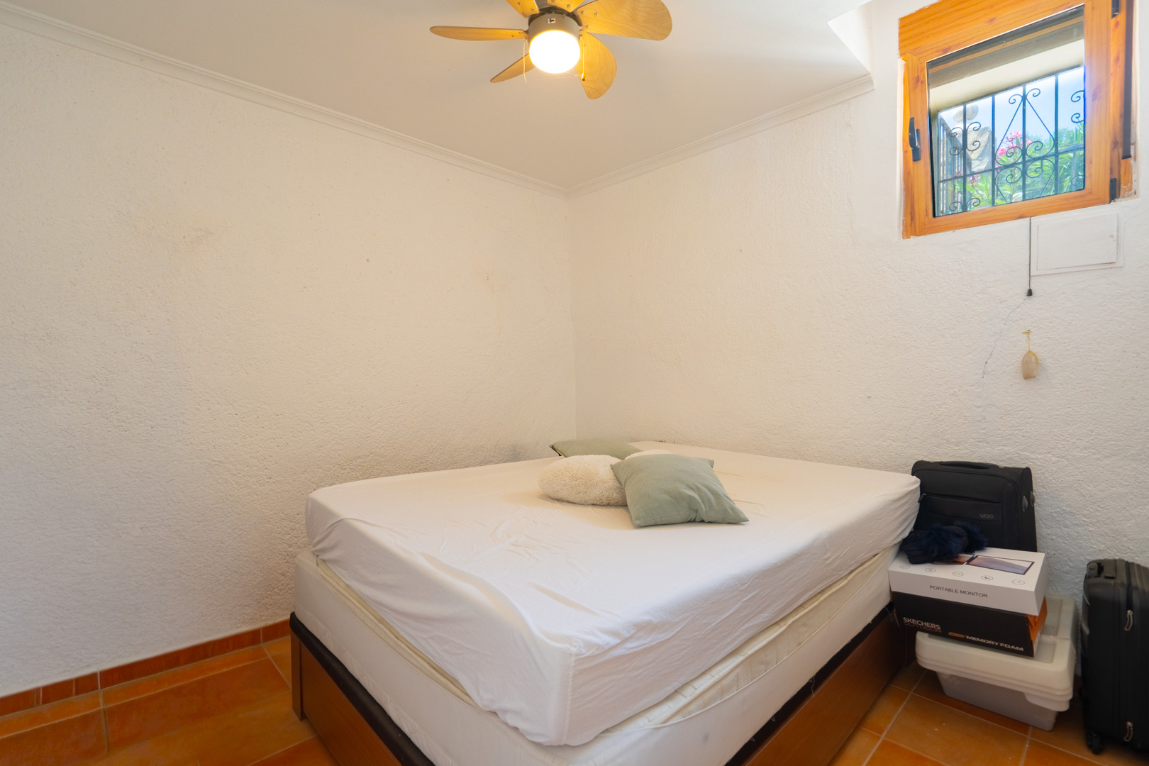 Dormitorio con cama doble, paredes blancas texturizadas y suelo de baldosas de terracota. Ventana de madera con rejas de seguridad y ventilador de techo. Dormitorio con cama doble, paredes blancas texturizadas y suelo de baldosas de terracota. Ventana de madera con rejas de seguridad y ventilador de techo.