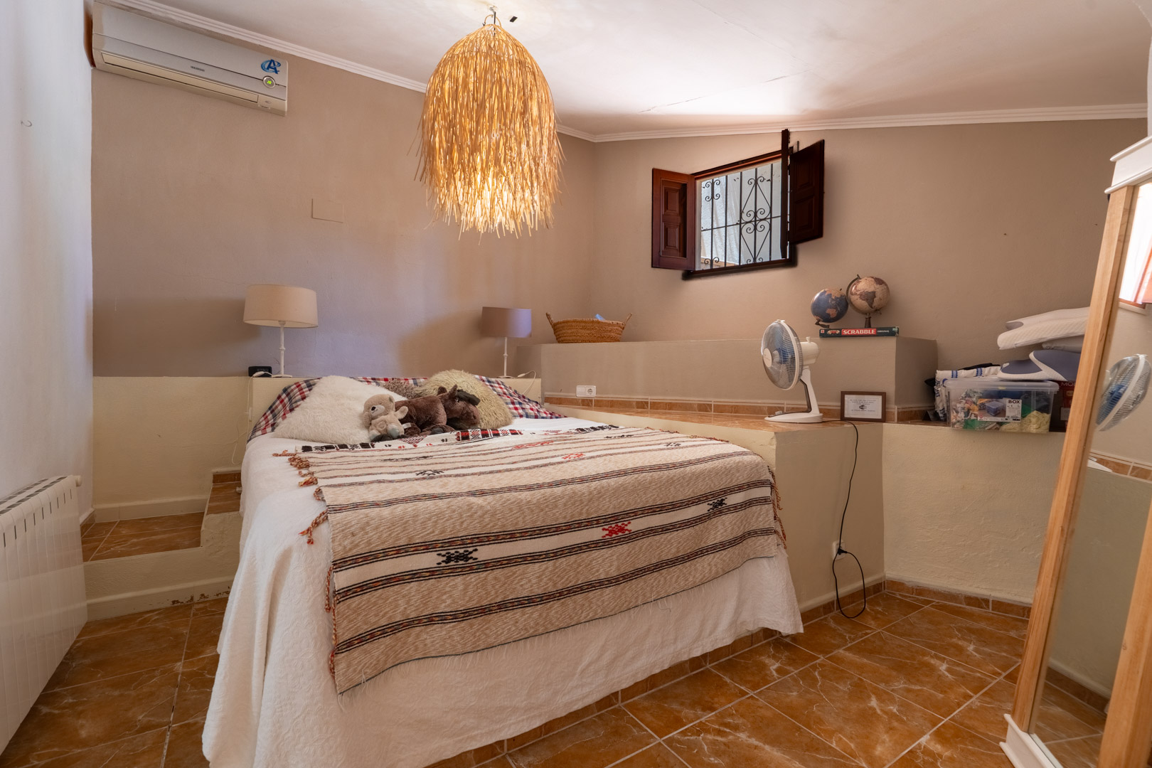 Dormitorio con cama doble sobre plataforma elevada, suelo de baldosas de terracota y lámpara colgante de mimbre. Ventana abierta con contraventanas de madera oscura y reja de hierro. Dormitorio con cama doble sobre plataforma elevada, suelo de baldosas de terracota y lámpara colgante de mimbre. Ventana abierta con contraventanas de madera oscura y reja de hierro.