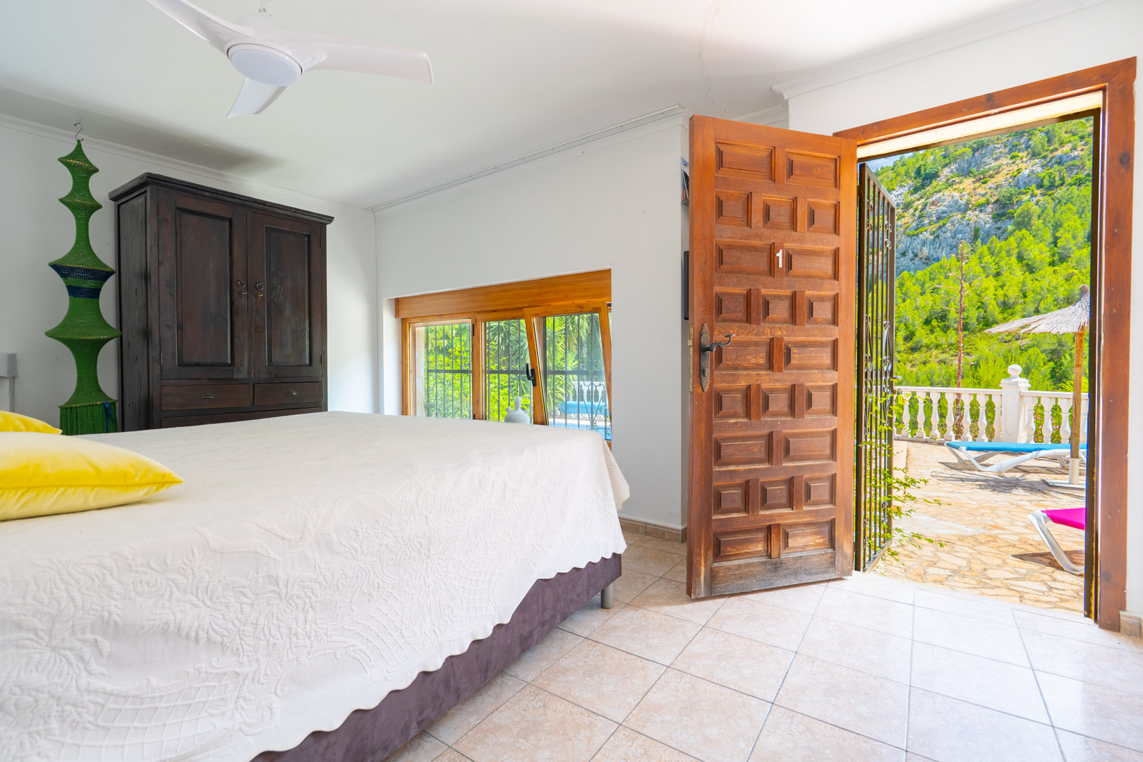 Dormitorio con suelo de baldosas claras, armario de madera oscura y ventilador de techo. Una puerta de madera abierta da a una terraza con vistas a la montaña y tumbonas. Dormitorio con suelo de baldosas claras, armario de madera oscura y ventilador de techo. Una puerta de madera abierta da a una terraza con vistas a la montaña y tumbonas.
