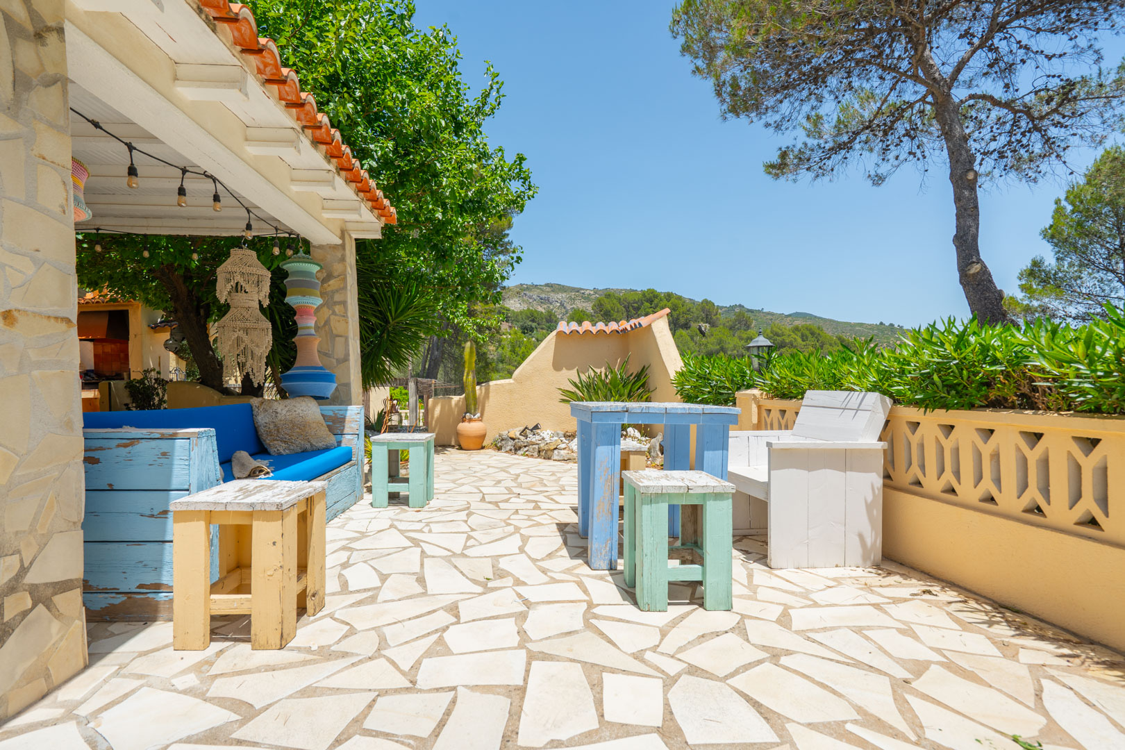 Encantador patio exterior con pavimento de piedra irregular, área de asientos cubierta con sofá azul y luces, junto a muebles de madera decapada y vegetación. Encantador patio exterior con pavimento de piedra irregular, área de asientos cubierta con sofá azul y luces, junto a muebles de madera decapada y vegetación.