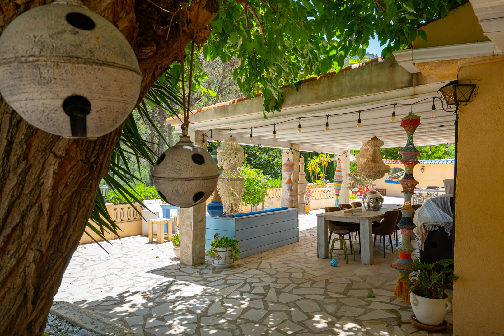 Encantadora zona de comedor exterior cubierta con mesa de madera rústica sobre pavimento de piedra, adornada con pilares artesanales y lámparas de cuentas, bajo un árbol frondoso con campanas. Encantadora zona de comedor exterior cubierta con mesa de madera rústica sobre pavimento de piedra, adornada con pilares artesanales y lámparas de cuentas, bajo un árbol frondoso con campanas.
