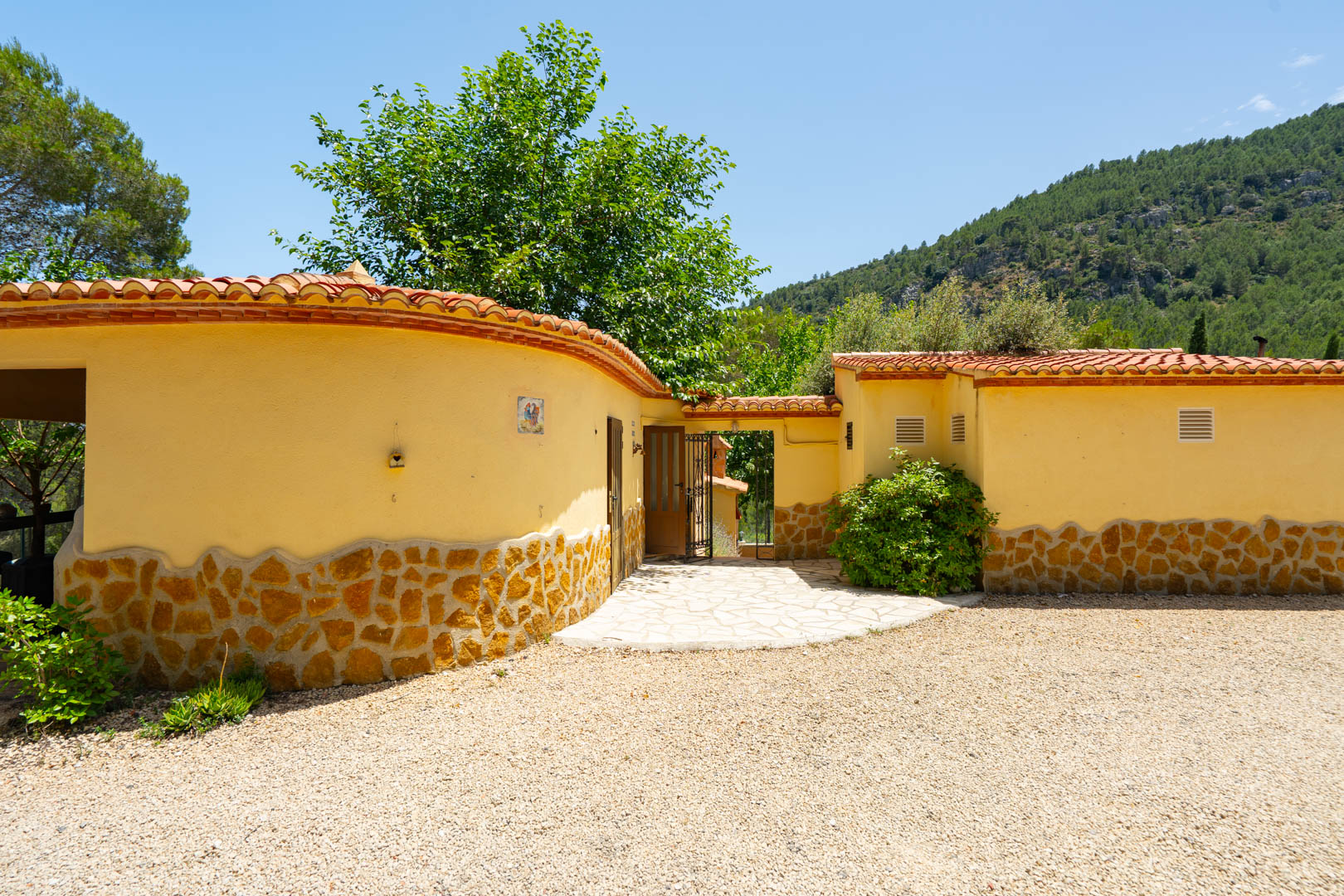 Entrada a la propiedad con camino de grava y sendero pavimentado, flanqueada por muros de estuco amarillo con zócalo de piedra natural y tejas de terracota, con fondo montañoso. Entrada a la propiedad con camino de grava y sendero pavimentado, flanqueada por muros de estuco amarillo con zócalo de piedra natural y tejas de terracota, con fondo montañoso.