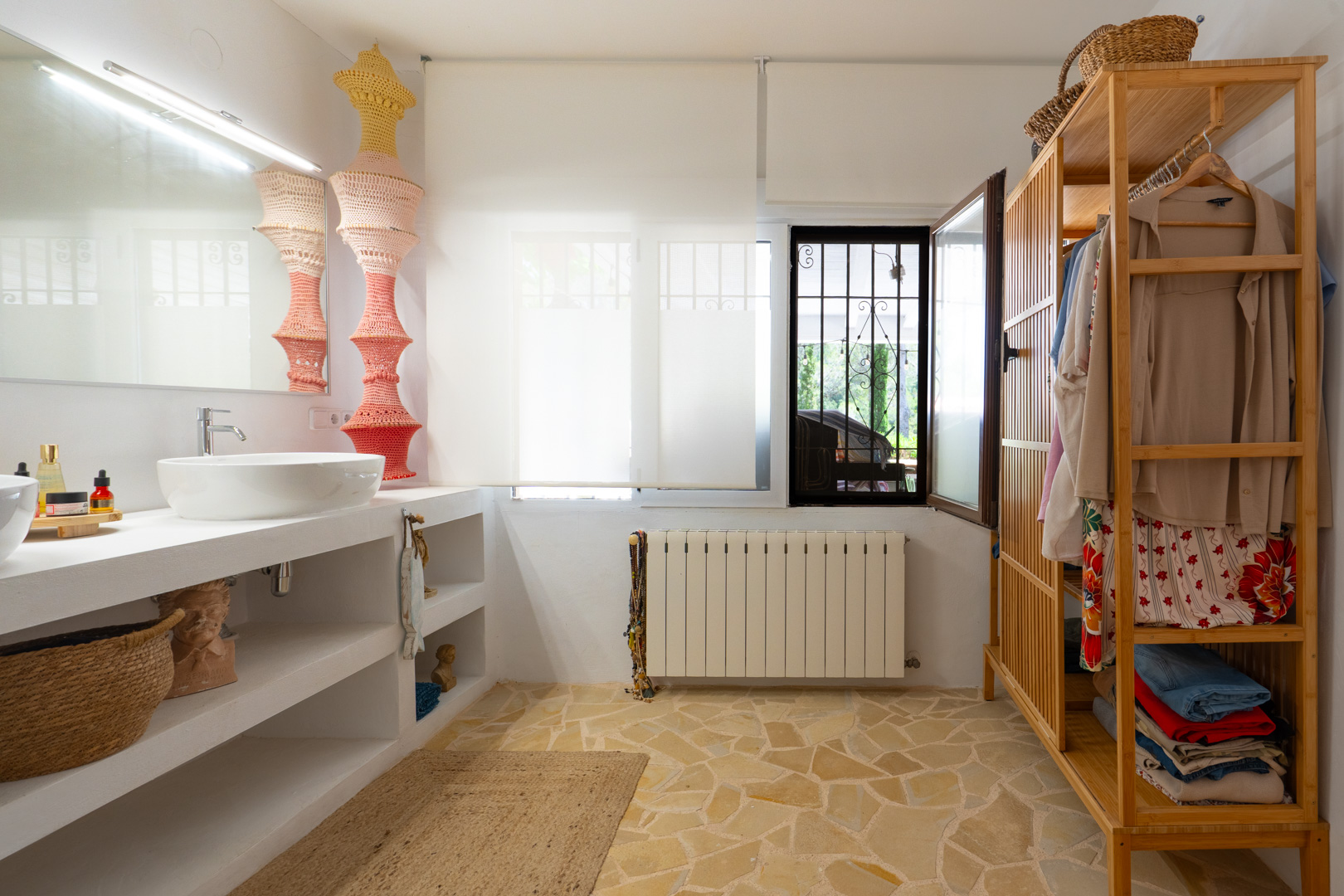 Espacio de baño funcional con lavabo sobre encimera blanca y estantes abiertos, sobre un suelo de baldosas de piedra irregular. Incluye un armario abierto de madera y ventana con reja de seguridad. Espacio de baño funcional con lavabo sobre encimera blanca y estantes abiertos, sobre un suelo de baldosas de piedra irregular. Incluye un armario abierto de madera y ventana con reja de seguridad.