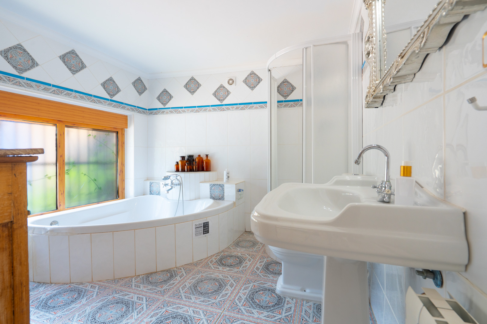 Este luminoso baño presenta una bañera esquinera alicatada bajo una ventana de madera, junto a un lavabo de pedestal y una cabina de ducha. Los azulejos estampados decoran el suelo y el borde de la pared. Este luminoso baño presenta una bañera esquinera alicatada bajo una ventana de madera, junto a un lavabo de pedestal y una cabina de ducha. Los azulejos estampados decoran el suelo y el borde de la pared.