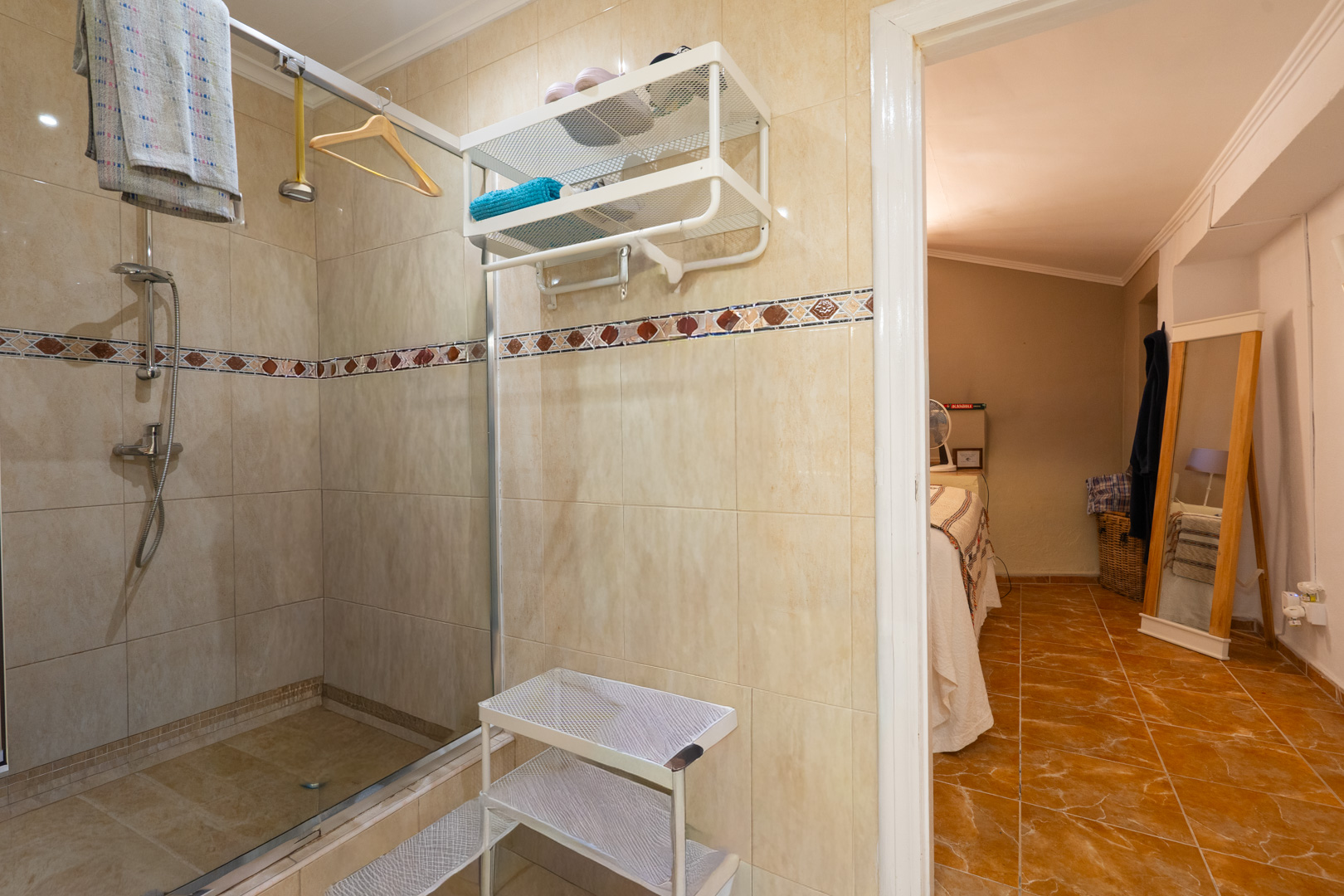 Funcional baño con ducha de azulejos beige, mampara de cristal y cenefa decorativa. Una puerta abierta revela un dormitorio adyacente con suelo de baldosas de terracota y espejo de cuerpo entero. Funcional baño con ducha de azulejos beige, mampara de cristal y cenefa decorativa. Una puerta abierta revela un dormitorio adyacente con suelo de baldosas de terracota y espejo de cuerpo entero.