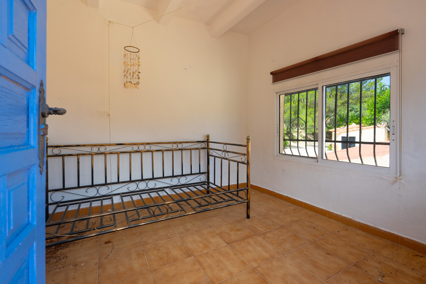 Habitación luminosa con un marco de diván de metal, suelo de baldosas de terracota y techo con vigas blancas. Una puerta azul se abre al espacio, y la ventana con rejas ofrece vistas a la vegetación. Habitación luminosa con un marco de diván de metal, suelo de baldosas de terracota y techo con vigas blancas. Una puerta azul se abre al espacio, y la ventana con rejas ofrece vistas a la vegetación.